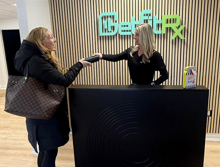 The real GetFitRx Cranston reception: logo on the wood wall, black desk, receptionist and customer